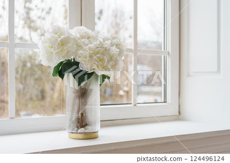Bouquet of white artificial decorative hydrangea, hortensia in vase on windowsill in living room 124496124