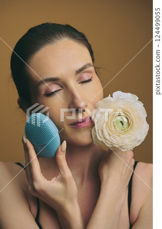 Woman using skincare device while holding a flower with closed eyes 124499055