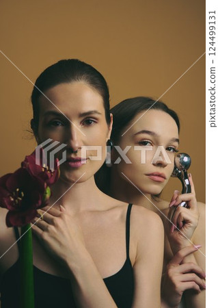 Two women pose with a flower and beauty tool against a warm backdrop Two women pose with a flower and beauty tool against a warm backdrop 124499131