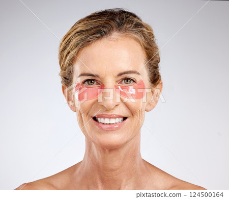 Face, beauty and antiaging with a senior woman using under eye patch cosmetics for skincare in studio on a gray background. Portrait, treatment and product with a mature female posing for wellness 124500164