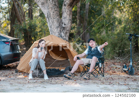 Two friends relax by a tent in a forest, enjoying a peaceful camping experience with a car and scooter nearby. 124502100