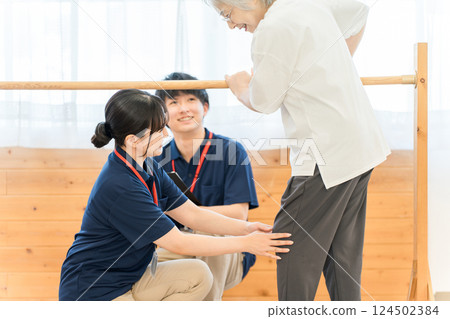 Senior woman using handrails for walking training and male and female occupational therapist and physiotherapist staff assisting her 124502384