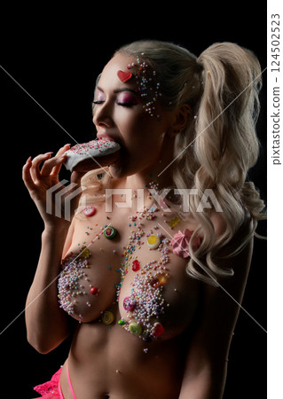 Woman with body art adorned in candy elements eating a dessert in a dark setting Woman with body art adorned in candy elements eating a dessert in a dark setting 124502523