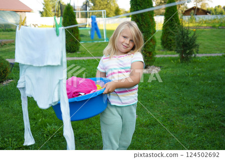 The girl, around six years old, waits near the clothesline with a plastic tub, gazing at the swaying sheets. 124502692