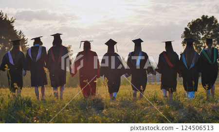 Group of graduates walking away in field at sunset holding hands, celebrating their academic achievement and friendship 124506513