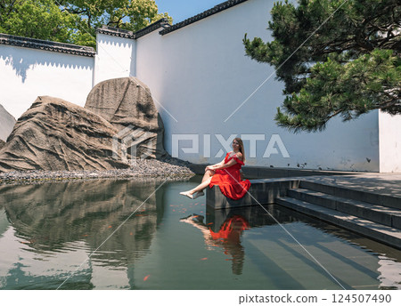 Woman in red dress relaxing by serene water in a tranquil garden in summer Woman in red dress relaxing by serene water in a tranquil garden in summer 124507490