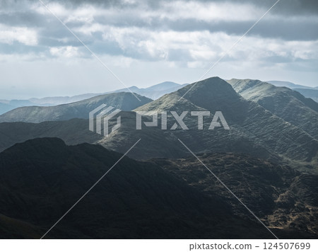 Overcast sky above a series of dark-colored mountains. The mountains display varying textures and slopes. The scene lacks bright light. Overcast sky above a series of dark-colored mountains. The mountains display varying textures and slopes. The scene lacks bright light. 124507699