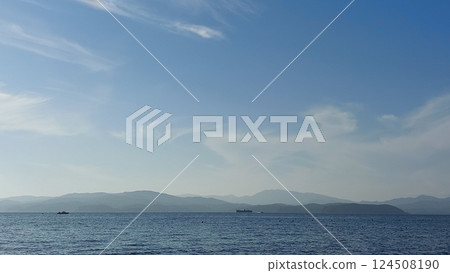 This photo captures a serene coastal scene. The image shows a vast, calm ocean extending into the horizon under a bright blue sky filled with scattered, fluffy white clouds. On the left side of the ph 124508190