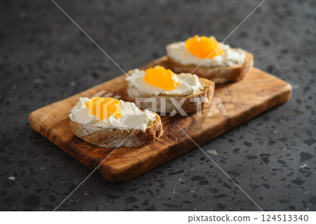Three ciabatta slices with ricotta and orange marmalade on olive wood board on terrazzo surface 124513340