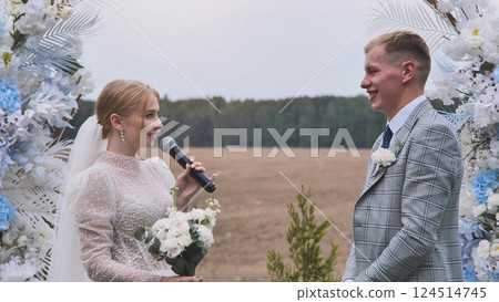 Bride holding microphone and giving a speech to groom during wedding ceremony under floral arch Bride holding microphone and giving a speech to groom during wedding ceremony under floral arch 124514745