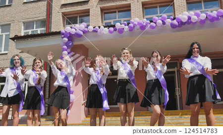 Cheerful female graduates waving goodbye on their last day of school, celebrating the end of the academic year with purple ribbons and balloons Cheerful female graduates waving goodbye on their last day of school, celebrating the end of the academic year with purple ribbons and balloons 124514783