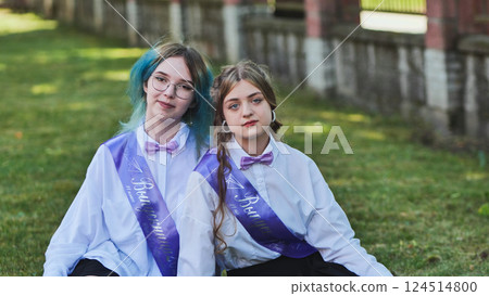 Two happy graduates celebrating their last school day, sitting on the grass in the schoolyard, wearing sashes and enjoying the moment as friends and classmates Two happy graduates celebrating their last school day, sitting on the grass in the schoolyard, wearing sashes and enjoying the moment as friends and classmates 124514800