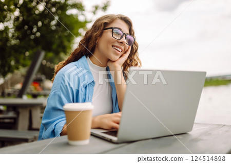 Portrait of a young woman in a blue shirt with a laptop at a table in an outdoor cafe near a lake. 124514898