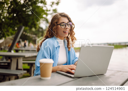 Portrait of a young woman in a blue shirt with a laptop at a table in an outdoor cafe near a lake. 124514901