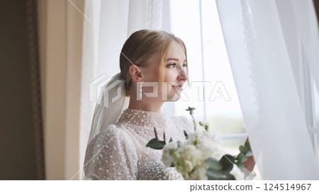 White clad bride holding wedding bouquet, gazing through window with soft, romantic expression White clad bride holding wedding bouquet, gazing through window with soft, romantic expression 124514967