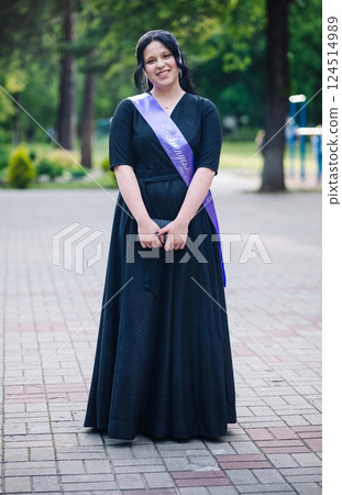 Young graduate student wearing a sash, smiling joyfully in a park, celebrating her recent graduation achievement surrounded by nature 124514989