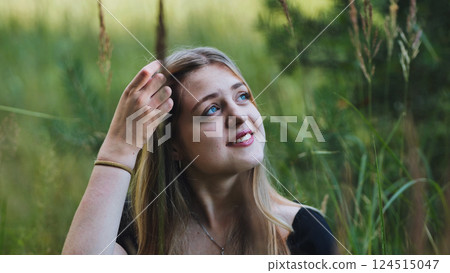Blonde teenager with radiant smile touching hair, standing amid tall green grass under bright sunlight 124515047