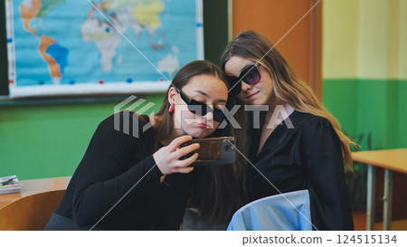 Two high school girls wearing sunglasses are using a smartphone in a classroom, possibly diverting their attention from their studies 124515134