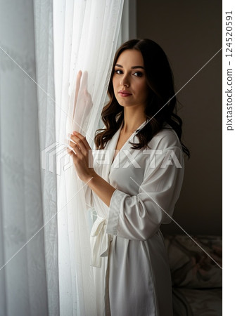 European brunette woman holding sheer white curtain with soft light in linen interior European brunette woman holding sheer white curtain with soft light in linen interior 124520591