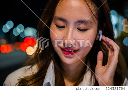 A smiling young woman using earphones attached to her mobile phone for a conversation on a night city street. Technology and wireless connection make it possible. Portrait of a happy girl. 124521764