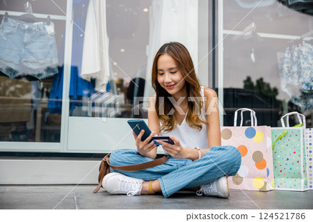 Young woman taking a break from shopping, sitting on the ground with bags, phone, and credit card. Online browsing and mobile payments make retail therapy a breeze Young woman taking a break from shopping, sitting on the ground with bags, phone, and credit card. Online browsing and mobile payments make retail therapy a breeze 124521786