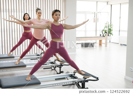 Three Asian women in pink sportswear doing pilates exercises with a reformer bed. Three Asian women in pink sportswear doing pilates exercises with a reformer bed. 124522692