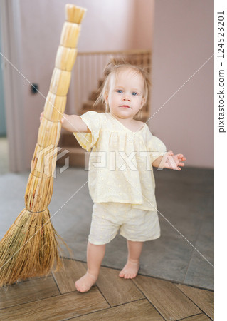 Full length vertical portrait of cute toddler girl standing barefoot on floor holding broom in modern living interior, helping with household chores and learning responsibility from young age. 124523201