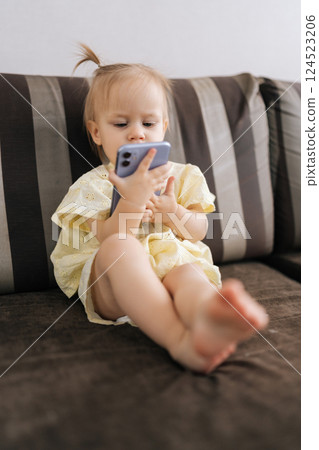 Vertical shot of focused toddler girl engaging with smartphone screen, exploring digital content while sitting comfortably at home, representing early technological interaction alone. 124523206