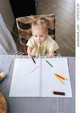 Vertical top view of little girl drawing with colored pencils on paper notebook, sitting at table, expressing creativity and enhancing fine motor skills while enjoying artistic play at home. 124523216