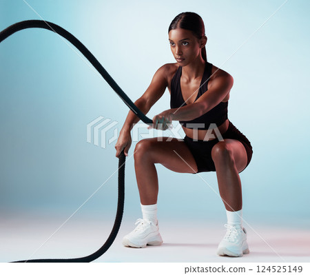Battle rope, fitness and woman in studio for exercise, strength training and cardio workout burning calories. Wellness, focus and healthy girl in a squat pose moving ropes with a blue background 124525149