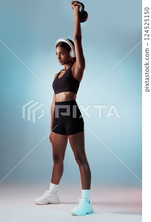 Health, exercise and woman in headphones with kettlebell on a blue studio background. Fitness, sports and female from India streaming music, radio or podcast while training with exercising weight. 124525150