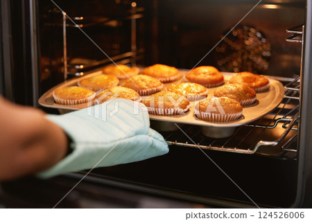 Hand, oven and muffins in baking, food or cooking sweet delicious cakes on a tray at home in the kitchen. Hands of baker taking hot muffin baked meal, treat or delight in pastry making at the house 124526006
