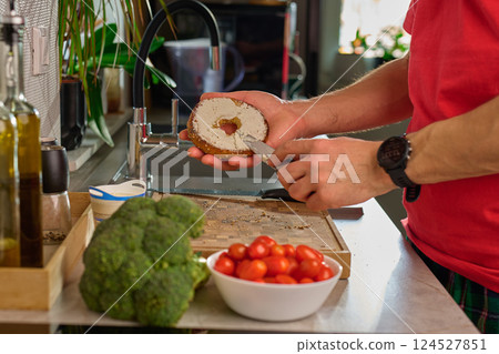 Man spreading cream cheese on bagel in kitchen, preparing sandwich 124527851