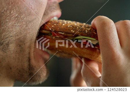 Mouth taking bite from crunchy bagel sandwich with greens and tomato 124528023