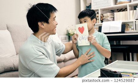 Happy son showing handmade Father Day card with red heart to smiling father in living room, bonding moment, camera and colored pencils on table, bookshelf in background Happy son showing handmade Father Day card with red heart to smiling father in living room, bonding moment, camera and colored pencils on table, bookshelf in background 124529887
