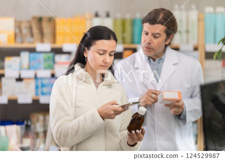 Middle-aged woman scanning QR-code on syrup while pharmacist offering Middle-aged woman scanning QR-code on syrup while pharmacist offering 124529987
