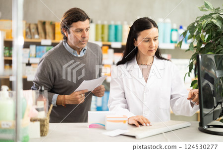 Middle-aged man standing with prescription and pharmacist looking to computer 124530277