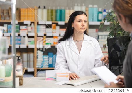 Female pharmacist standing behind counter with computer and talking to man 124530449