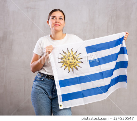 Joyful young female holding national flag of Uruguay while standing against gray wall 124530547