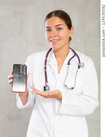 Female doctor in white uniform recommending mobile medical app, stretching out hand with smartphone. Focus on device screen 124530592