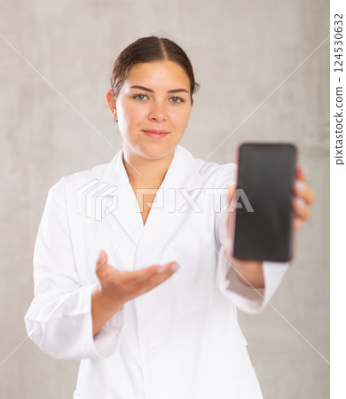 Female doctor in coat extending hand with phone and showing screen, selective focus on gadget. Online consultation concept Female doctor in coat extending hand with phone and showing screen, selective focus on gadget. Online consultation concept 124530632