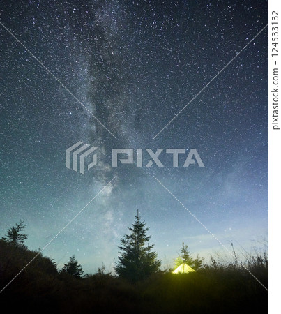 Radiant yellow tent beneath awe-inspiring, star-filled sky with Milky Way prominently stretching across. Surrounded by pine trees and silhouetted by hill, scene epitomizes tranquility of nature. Radiant yellow tent beneath awe-inspiring, star-filled sky with Milky Way prominently stretching across. Surrounded by pine trees and silhouetted by hill, scene epitomizes tranquility of nature. 124533132