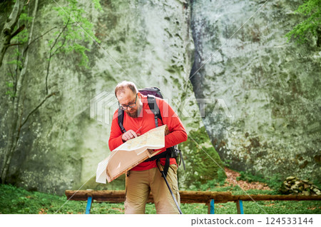 Bearded tourist man looks puzzled, studies map while stands in front of large rock in dense forest. Traveler with grey backpack and glasses, trying to figure out his direction. 124533144