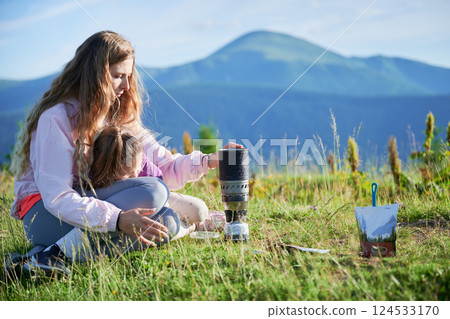 Woman prepares meal from packaged pouch next to portable camping stove. Female tourist with long blonde hair, sitting on grassy hillside with mountains in background, enjoying scenic outdoor setting. Woman prepares meal from packaged pouch next to portable camping stove. Female tourist with long blonde hair, sitting on grassy hillside with mountains in background, enjoying scenic outdoor setting. 124533170