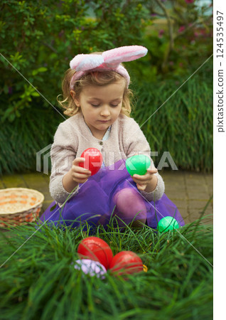 Easter Egg Hunt with Toddler. girl wearing bunny ears picks colorful Easter eggs hidden in the grass during an outdoor egg hunt, surrounded by greenery and springtime joy 124535497
