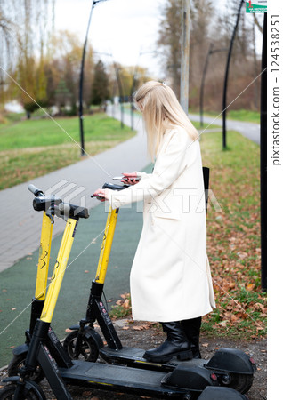 Elegant lady in a white coat checking her phone while standing by shared scooters on a tranquil park pathway during a crisp autumn day 124538251