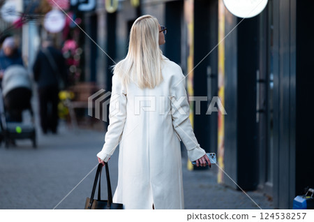 Walking through a vibrant street, a woman in a stylish white coat carries a bag while enjoying the atmosphere of a bustling city on a sunny day 124538257