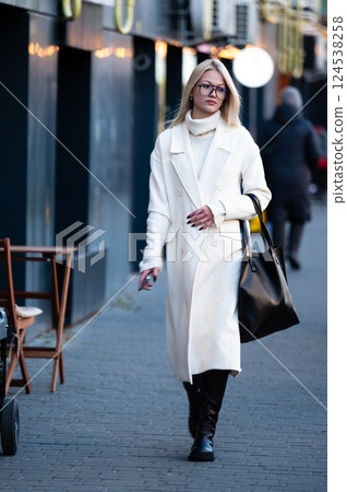 Stylish woman strolling through a city street in winter, wearing a chic white coat and black boots while carrying a large handbag on a chilly afternoon Stylish woman strolling through a city street in winter, wearing a chic white coat and black boots while carrying a large handbag on a chilly afternoon 124538258