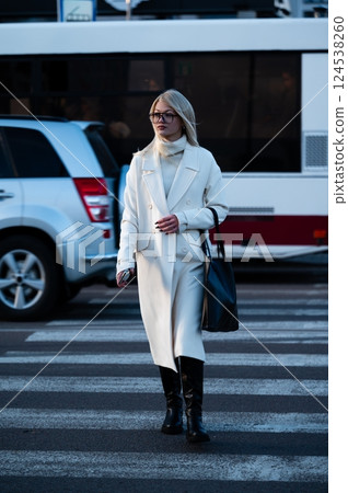 Walking confidently in the city, a fashionable woman in a long white coat crosses the crosswalk as vehicles await her stylish presence during a vibrant late afternoon Walking confidently in the city, a fashionable woman in a long white coat crosses the crosswalk as vehicles await her stylish presence during a vibrant late afternoon 124538260
