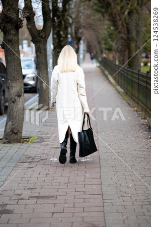 A stylish individual walks confidently along a tree-lined sidewalk, carrying a sleek black bag on a crisp autumn day while leaves rustle gently in the breeze A stylish individual walks confidently along a tree-lined sidewalk, carrying a sleek black bag on a crisp autumn day while leaves rustle gently in the breeze 124538269